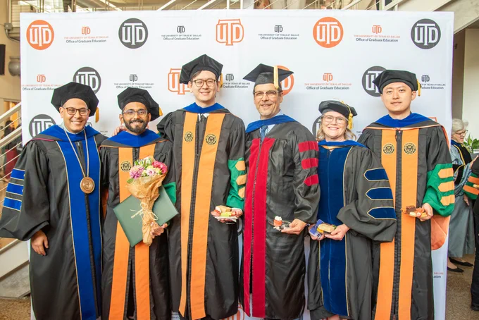 A group of six graduates in academic robes and caps smiles in front of a "UTD Office of Graduate Education" backdrop. One holds flowers, others hold small items.