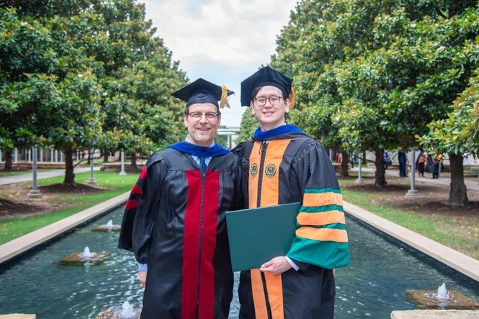 Dr. Schmidtke and Dr. Tjahjono in academic gowns and caps stand next to a fountain surrounded by lush green trees. They smile proudly, one holding a diploma.
