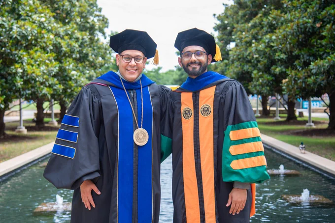 Dr. Obaid and Dr. Shah, in academic gowns and caps, stand next to a fountain surrounded by lush green trees. They smile proudly.