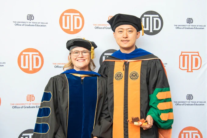 Dr. Jones and Dr. Shao, in academic regalia, pose smiling at a graduation event. A backdrop displays "UTD" and "Office of Graduate Education" logos.
