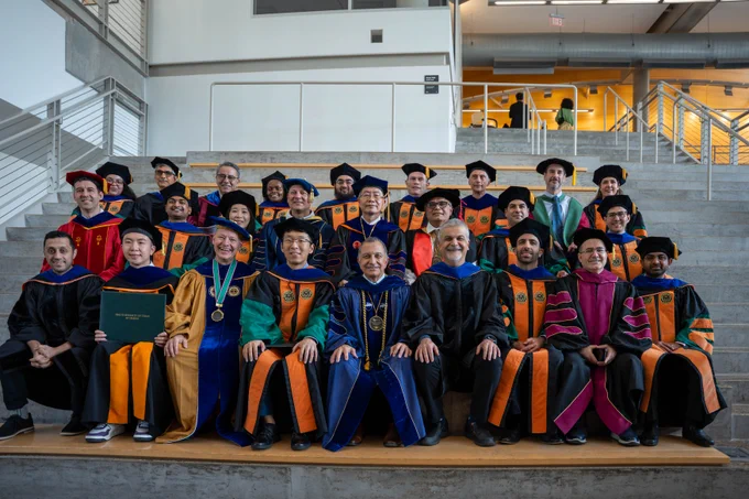 A group of people wearing academic regalia are seated on steps inside a building. They are dressed in robes and mortarboards of varying colors, indicating different academic disciplines. The setting is of a graduation ceremony.