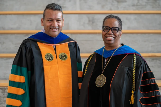 Two individuals wearing graduation robes stand side by side. They both have academic medallions and colored hoods.