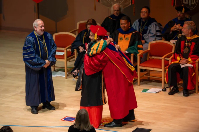 A person in academic regalia hugs another individual dressed in vibrant red robes on stage during a graduation ceremony. Several people in similar formal attire are seated in the background, observing the event.
