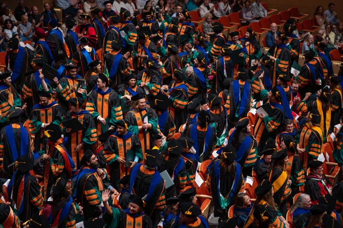 A large group of people dressed in graduation gowns and caps are gathered in an auditorium. Colorful academic hoods and robes are visible among the crowd, indicating a formal ceremony such as a graduation.