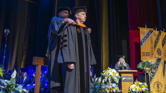 Tim Cahill stands on a platform on the commencement stage at Elliott Hall of Music as a Purdue official places an orange doctoral hood over his shoulders, with Purdue banners and flower arrangements visible in the background.