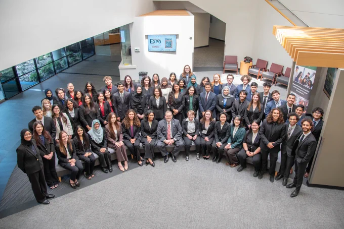 A large, diverse group of professionals in business attire gather in a spacious, modern lobby. They smile, conveying a sense of teamwork and achievement.