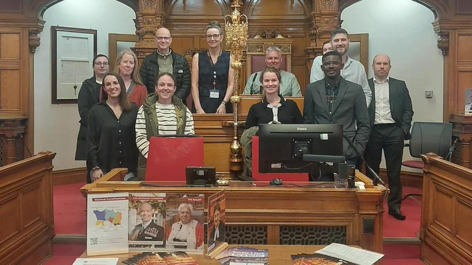Staff from the Government of Jersey. They are standing around the Bailiff's desk. The Royal Mace is in the centre. 