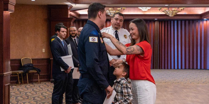 A person in a dark uniform is being honored by another person is placing a pin on the uniform while a small child stands nearby. 
