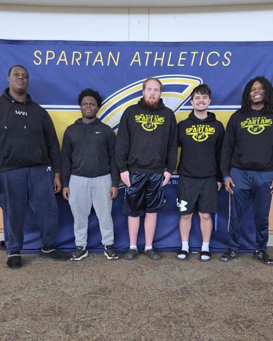 Five people standing in front of a "Spartan Athletics" banner. They are wearing casual athletic clothing, mostly consisting of hoodies and shorts.