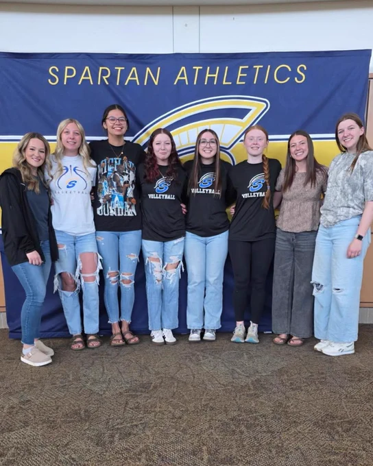 A group of people standing in front of a Spartan Athletics banner. They are dressed in casual and sports attire, with some wearing jeans and T-shirts, while others have sweatshirts and sneakers.