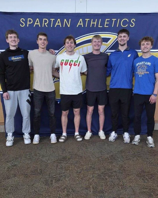 Six people standing in front of a "Spartan Athletics" banner, smiling and posing for a group photo. One person is wearing a Gucci shirt.