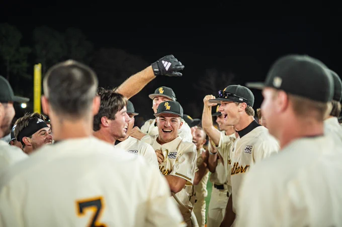 The Southern Miss baseball team, wearing cream jerseys and black baseball caps, huddles together cheering and throwing their arms into the air after winning a game.