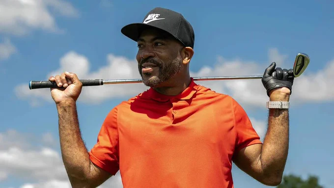A person wearing a red polo shirt and a black Nike cap is holding a golf club behind their shoulders, standing outdoors under a partly cloudy sky.