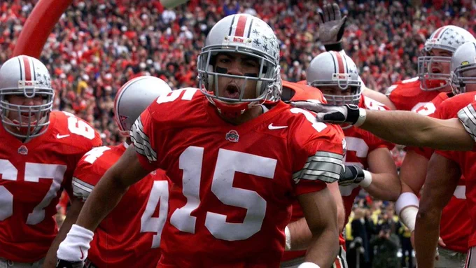 A football player wearing a red Ohio State jersey with the number 15 is on the field, surrounded by teammates. A large crowd is visible in the stadium background.