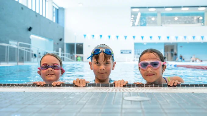 Three children wearing swim goggles hold onto the edge of an indoor swimming pool, ready for a swim session.