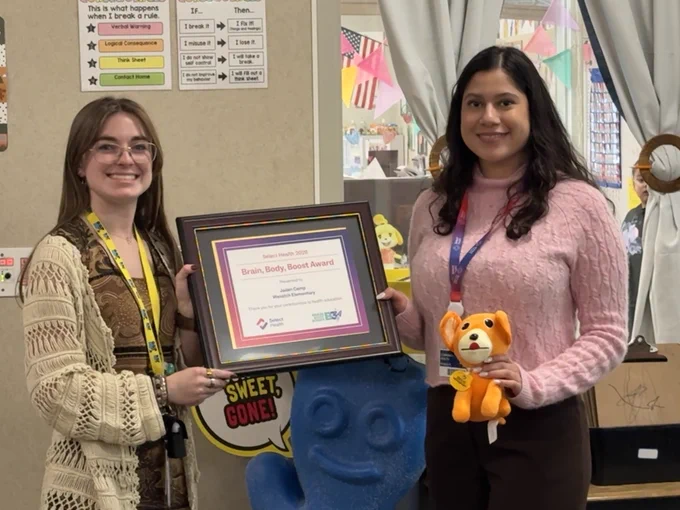 Two people stand in a room holding objects. One holds a framed certificate titled "Brain, Body, Boost Award" from Action for Healthy Kids, the other holds a small plush toy resembling a fox. Behind them are colorful decorations and educational materials on the walls.