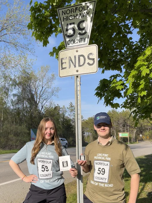 Two Ontario Parks employees model shirts that say, "Long Point Provincial Park End of the Road", with a Norfolk County 59 road sign in the middle of the shirt. One employee is wearing a green shirt and the other is wearing a blue shirt. They are standing just outside the park entrance, in front of the actual road sign that says, "Norfolk County Highway 59 Ends". 
