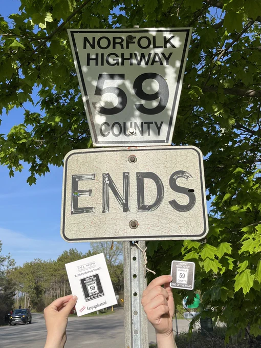 A sticker and a patch that say "Long Point Provincial Park End of the Road" are being held up to the "Norfolk County Highway 59" road sign that matches the sticker and patch.