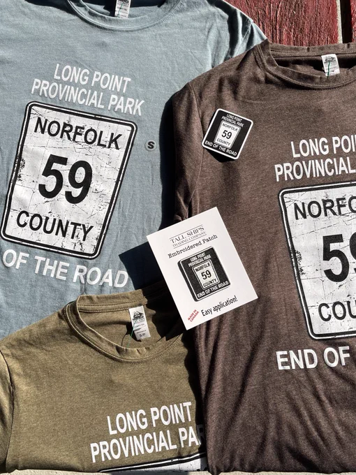All three colours (light blue, brown, and olive green) of the "Long Point Provincial Park End of the Road Highway 59" shirt are displayed on a picnic table with the matching sticker and patch laid out on top of the shirts.
