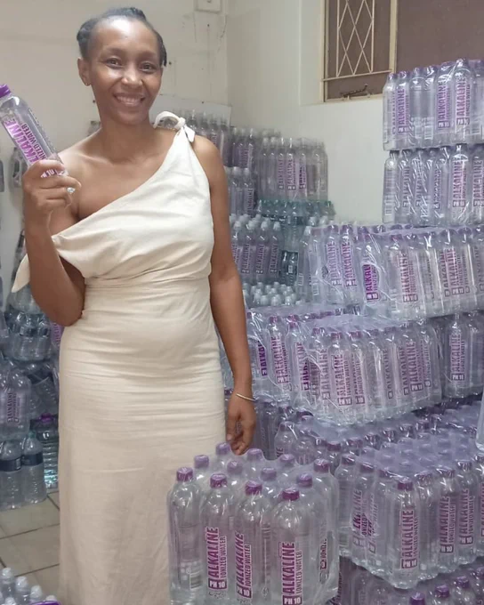 Nosisa holding a bottle of Designer Water pH10, standing next to stacks of cases of Designer Water pH10 in a warehouse in Pietermaritzburg