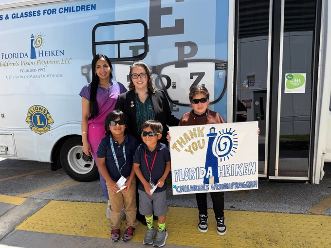 A group photo of two adults and three children smiling for the camera in front of the Florida Heiken Mobile Eye Care Unit. One child is holding a sign that reads "Thank you Florida Heiken Children's Vision Program"
