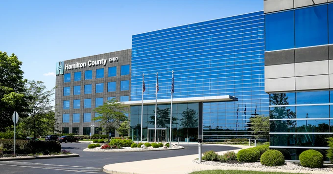 Modern office building with "Hamilton County Ohio" signage, glass facade, three flagpoles out front, landscaped greenery, and a clear blue sky.