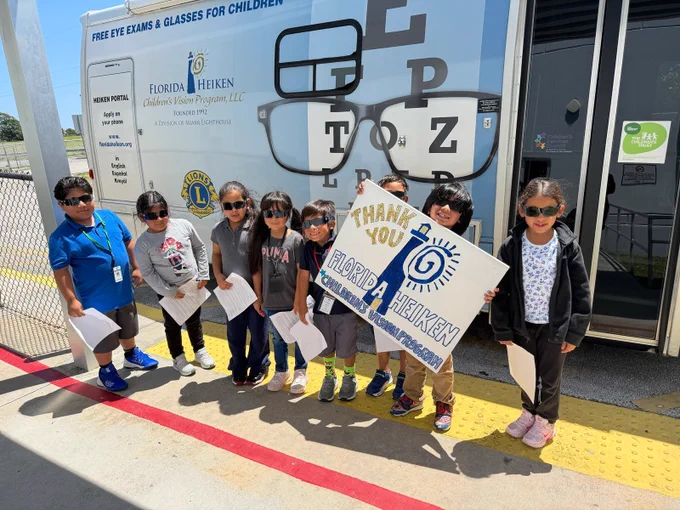 A group photo of 8 children wearing dilation sunglasses, smiling for the camera in front of the Florida Heiken Mobile Eye Care Unit. One child is holding a sign that reads "Thank you Florida Heiken Children's Vision Program".