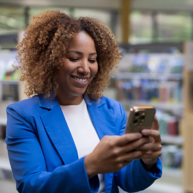 Photo of an individual using a smart phone in a library setting. There are bookshelves in the background and they are wearing a blue blazer and white top. 