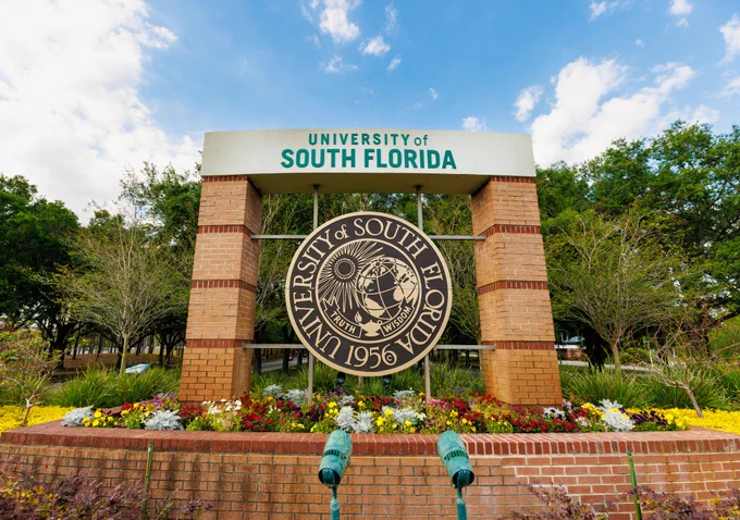 Signage with the University of South Florida seal, set against a backdrop of flowers, green trees, and a blue sky.