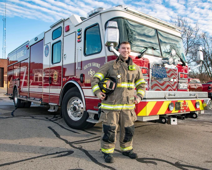 Gateway Fire Science student in full gear holding helmet stands confidently in front of a red fire truck on a clear day.