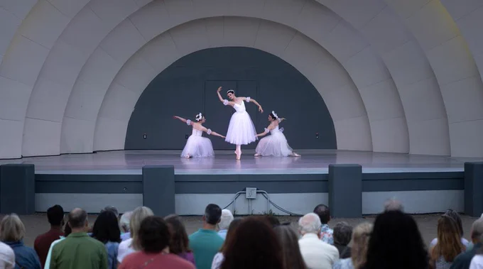 Three ballerinas in white tutus perform a graceful dance on an outdoor stage with an arched backdrop before an attentive audience.