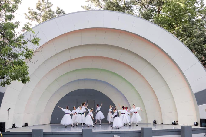 Ballet dancers in white tutus performing on an outdoor stage with a large, curved, rainbow-colored backdrop.