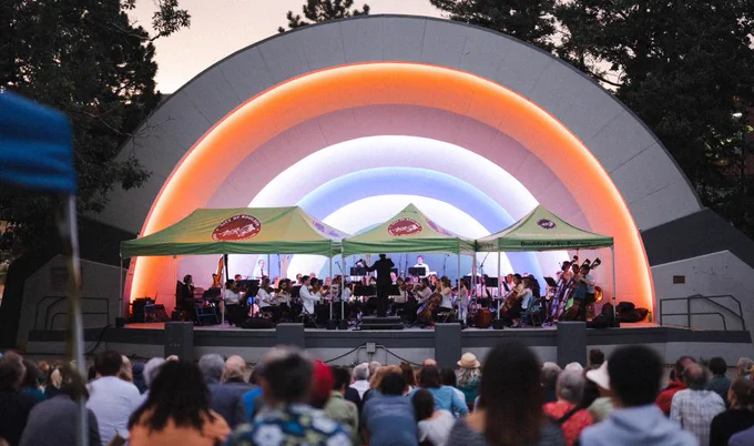 Outdoor orchestra performance under a large, illuminated rainbow-shaped stage arch at dusk with an attentive audience.