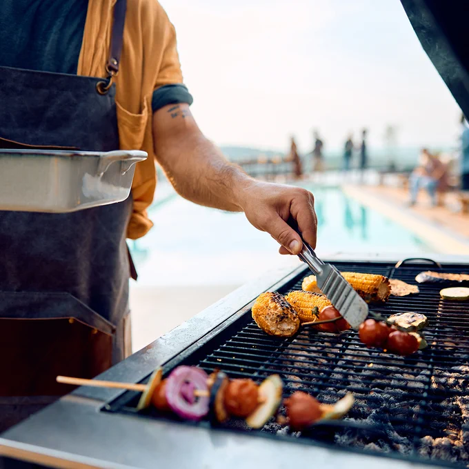 A person cooking vegetables and skewers on a grill near a poolside. Corn and other vegetables are visible on the grill. The background shows blurred figures by the pool.