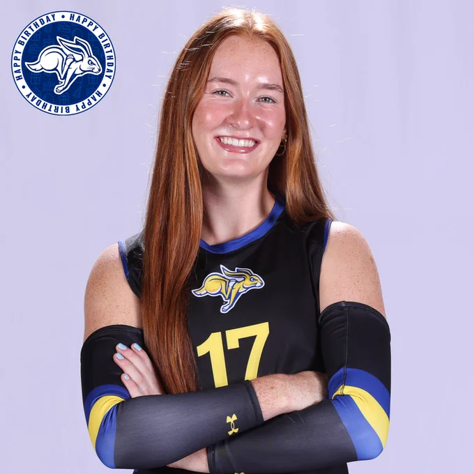 Studio portrait against a light gray background showing an athlete with long red hair, arms crossed, wearing a black South Dakota State uniform with blue and yellow accents and the number 17 on the front. The uniform includes arm sleeves with blue and yellow stripes. A Jackrabbit logo is centered on the chest. In the top left corner, a circular blue graphic with a Jackrabbit logo repeats the words “Happy Birthday.”
