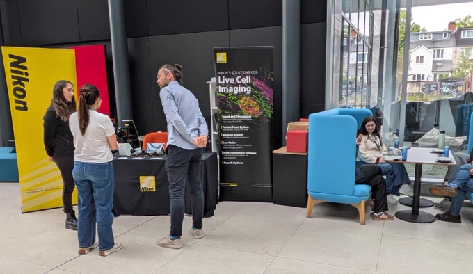 Three people engage in conversation at a Nikon booth featuring live cell imaging displays in a modern indoor space.