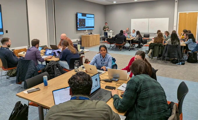 A group of people collaborating in a modern conference room with laptops and a presenter at the front near a screen and whiteboards.