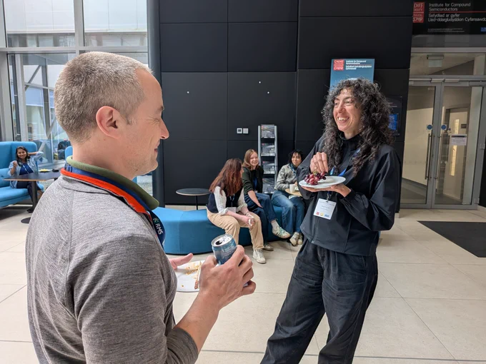 Two people engaged in conversation during a casual indoor event, one holding a drink and the other eating from a plate.
