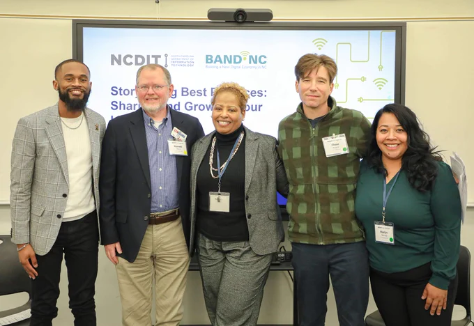 Five professionals standing together in front of a presentation screen displaying NCDIT and BAND-NC logos at a conference.