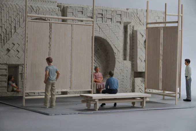 Architectural model with miniature figures in a scene featuring a textured stone wall and fabric screens.