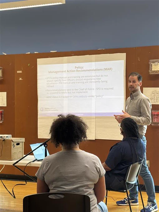Picture of OPA’s policy manager, Justin Piccorelli, presenting at the Human Rights Commission. He is wearing a gray shirt and blue jeans standing next to a screen presentation that says “Policy: Management Action Recommendations (MAR).” In front of Justin are two people sitting with their backs to the camera, but facing the screen. One is in a gray t-shirt with their dark hair in a ponytail. The other is closer to Justin in a dark blue polo with dark hair in braids. 