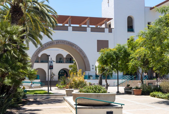 The image shows the Conrad Prebys Aztec Student Union at San Diego State University. The building features a white facade with a prominent archway and mission-style architecture. Palm trees and plants decorate the surrounding plaza area.