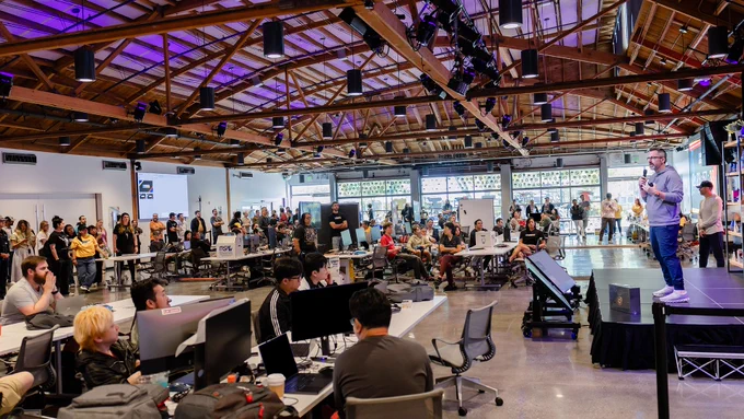 Large group of Rioters gathered in the Thunderdome event space with rows of computers, stage lighting, and a Rioter speaking onstage to the crowd beneath exposed wooden beams.
