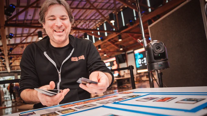 Rioter smiling while holding Riftbound trading cards over a taped tabletop set up Thunderdome.
