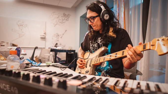 Rioter wearing headphones playing an electric guitar beside a keyboard and music equipment during Thunderdome in a music studio-style workspace.
