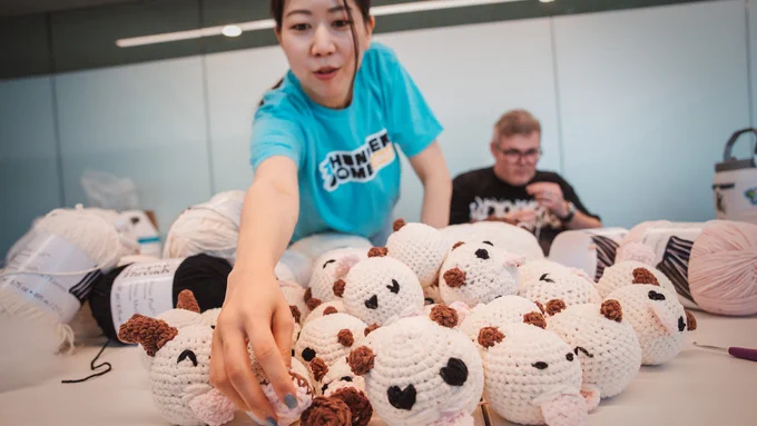 Rioter reaching across a table filled with crocheted white poro plushies and yarn supplies during Thunderdome, with another Rioter seated in the background.

