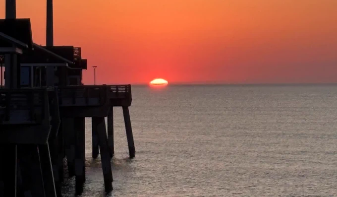 Sunrise and pier, with the sun partially below the horizon casting a warm orange glow over the calm ocean.