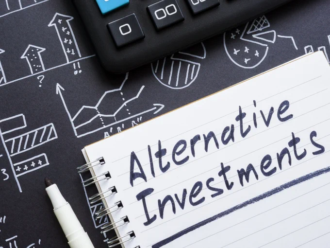 The word Alternative Investments is written on notebook paper with a calculator and hand-drawn charts on the desktop.