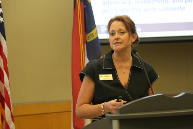 Woman in black top speaking at a podium with American and state flags behind her in a conference room.