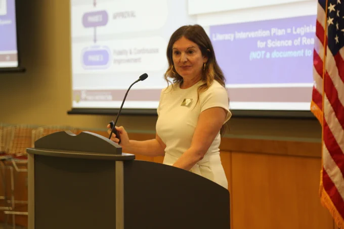 Woman in white dress speaking at a podium with a presentation slide and American flag in the background.